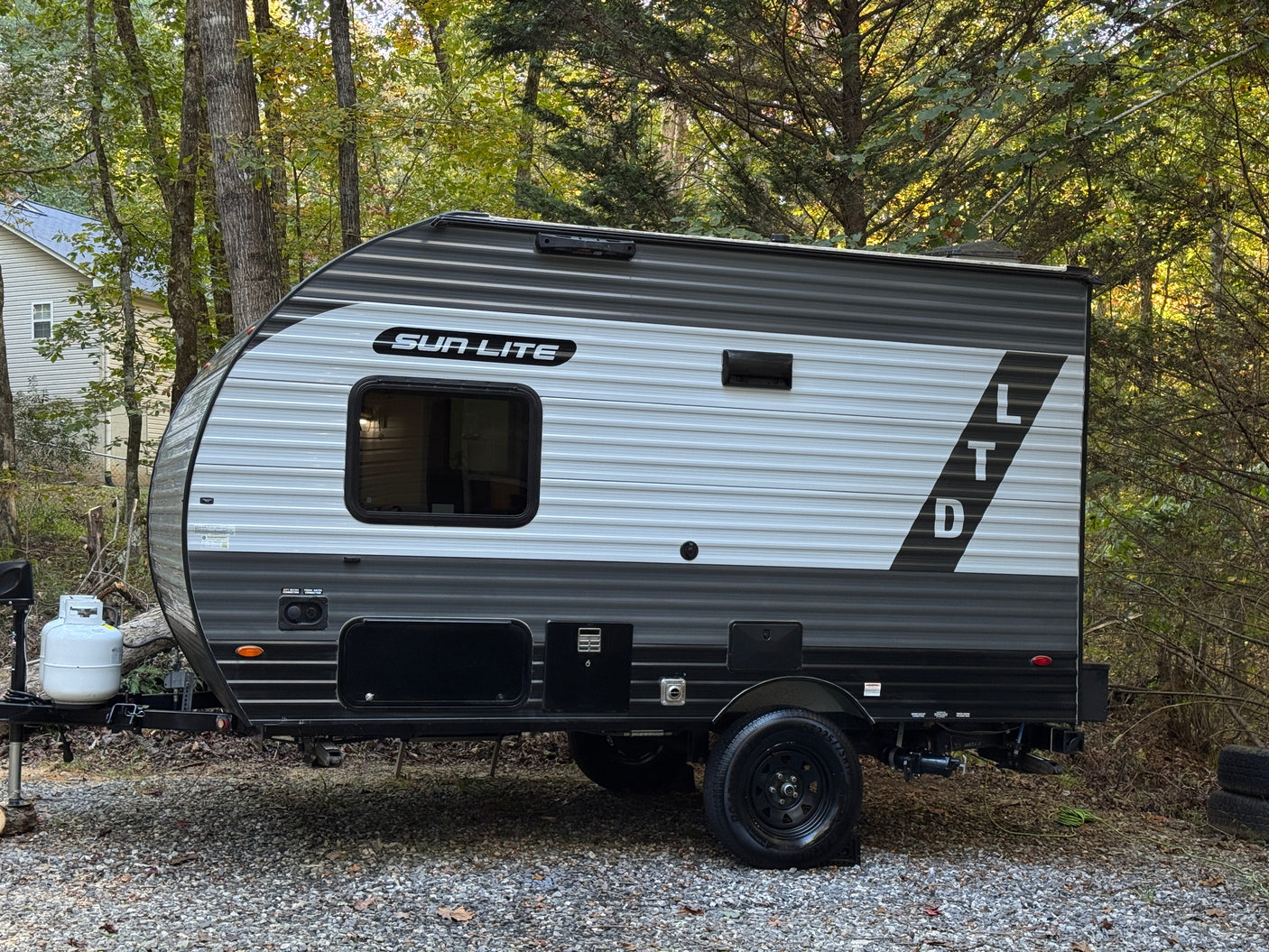 Grey and white Sun Lite LTD travel trailer, an ideal RV rental, parked on a gravel lot surrounded by lush trees.