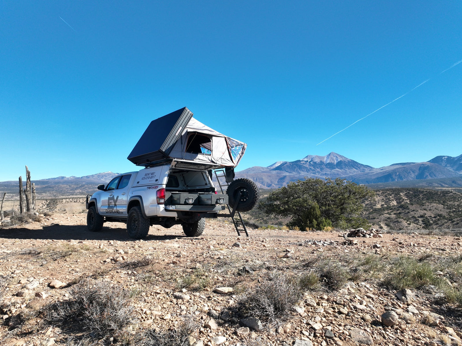 Silver overlanding RV rental truck with a rooftop tent deployed, parked in a rocky desert landscape with mountains.