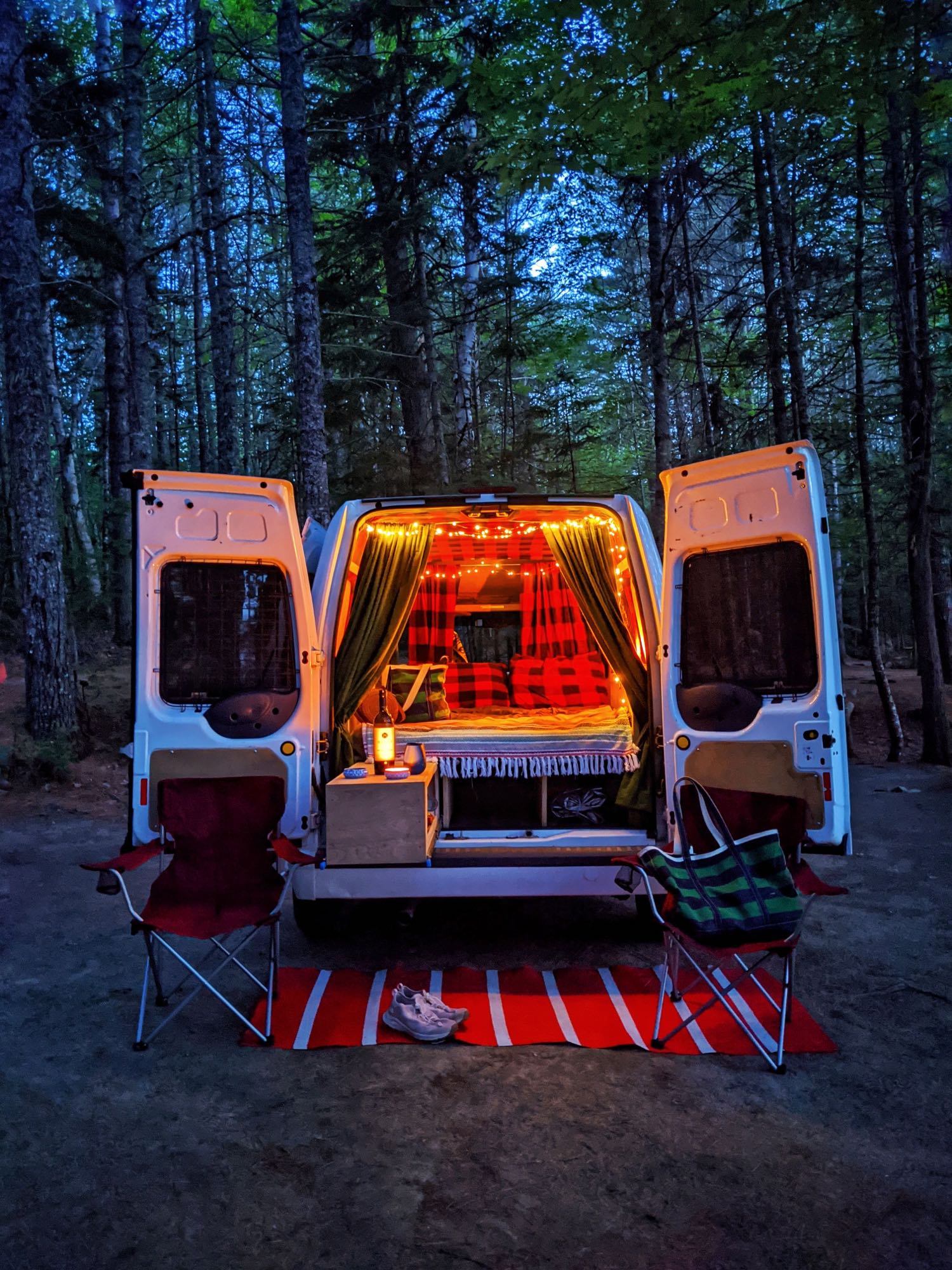 Cozy RV rental adventure: A camper van with glowing lights and open doors in a dark forest. Chairs & a rug sit outside.|White "Maine Campervan Rentals" van