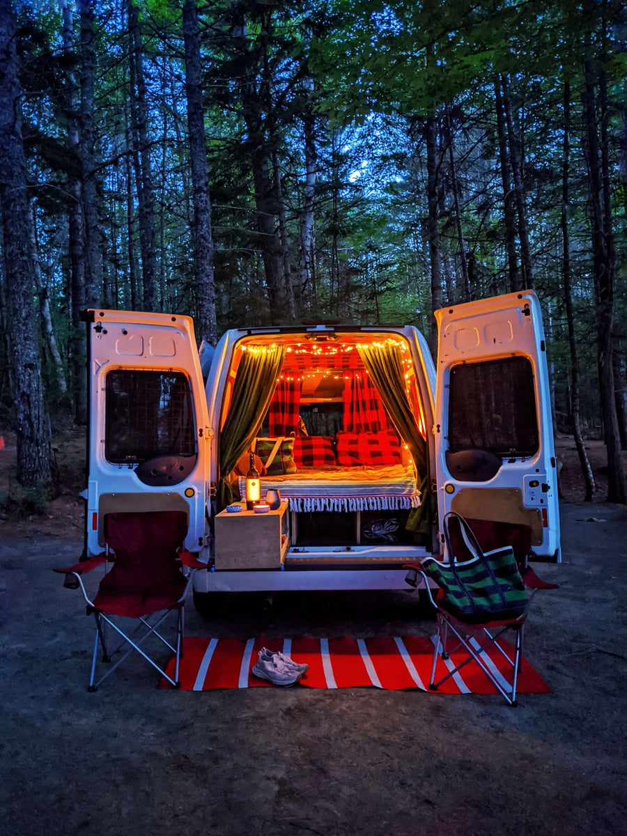 Cozy RV rental adventure: A camper van with glowing lights and open doors in a dark forest. Chairs & a rug sit outside.|White "Maine Campervan Rentals" van