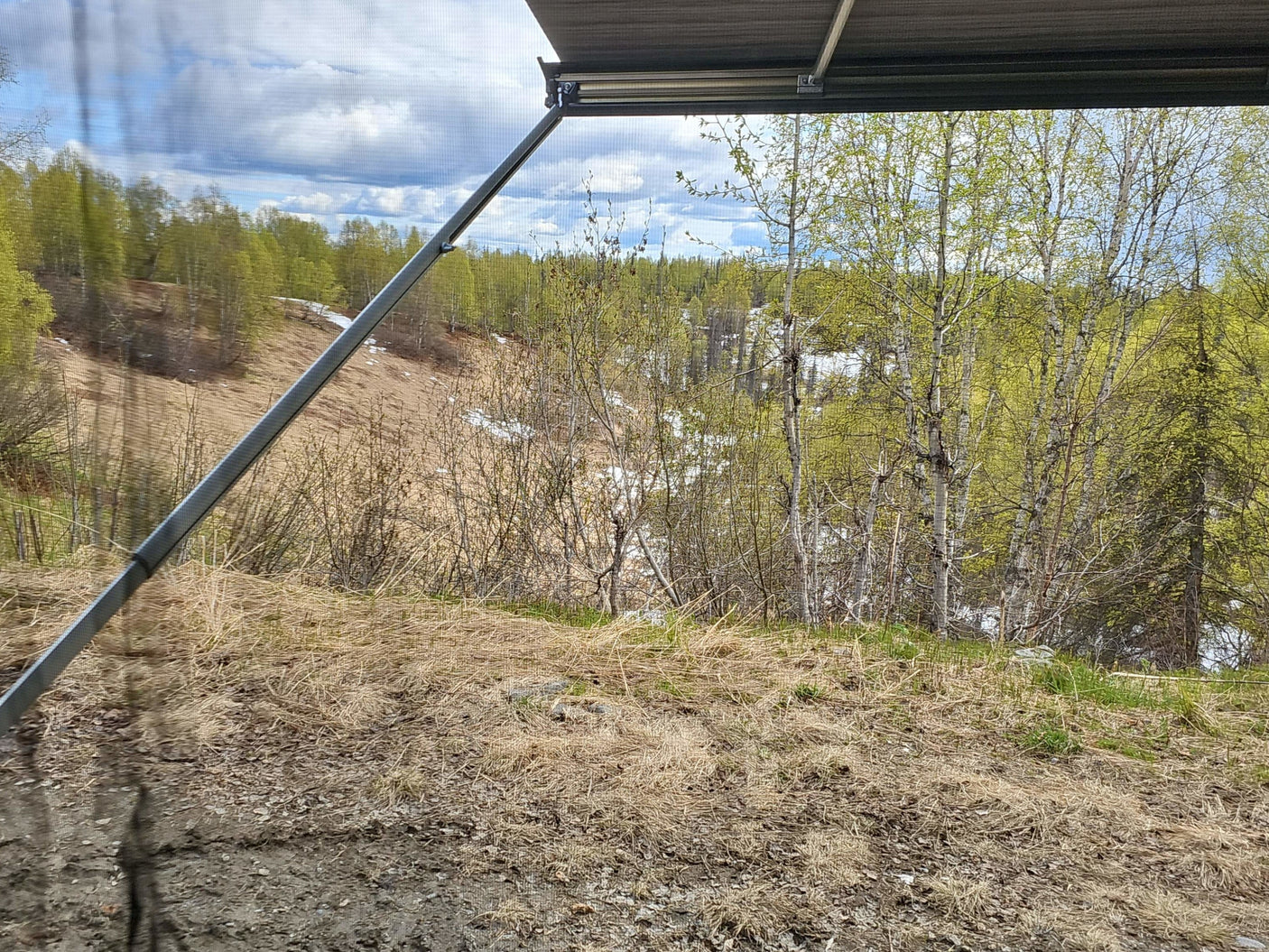 Scenic spring view from an RV rental. Through screen, green trees, melting snow, and cloudy sky are visible.