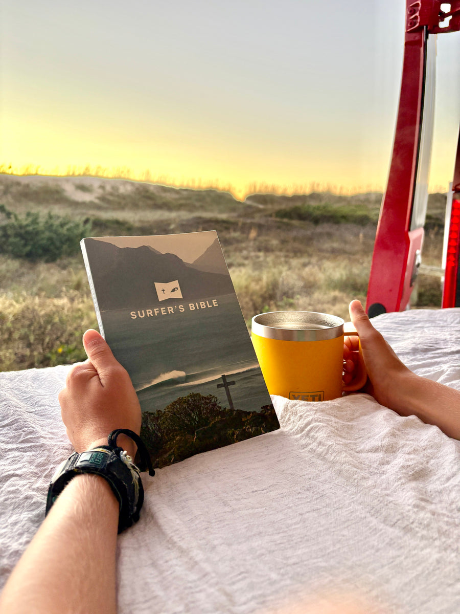Enjoying an **rv rental** view: hands holding 'Surfer's Bible' and YETI mug, overlooking beach dunes at sunset/sunrise. A quiet spiritual retreat.
