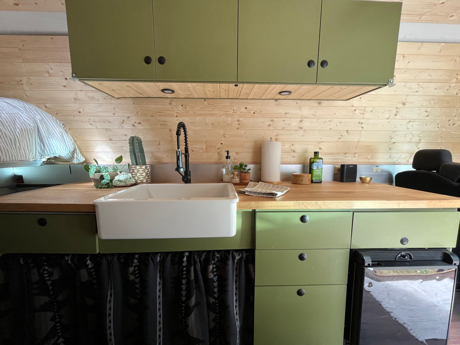 Compact RV rental kitchen with olive green cabinets, wooden walls, a white sink, and wood countertop.