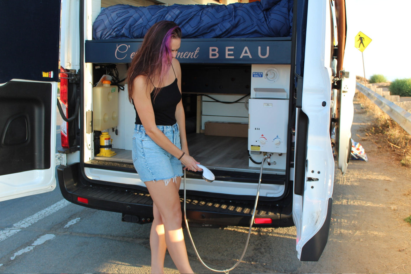 Woman showering at rear of RV rental van, showcasing outdoor shower system with portable water heater and bed.