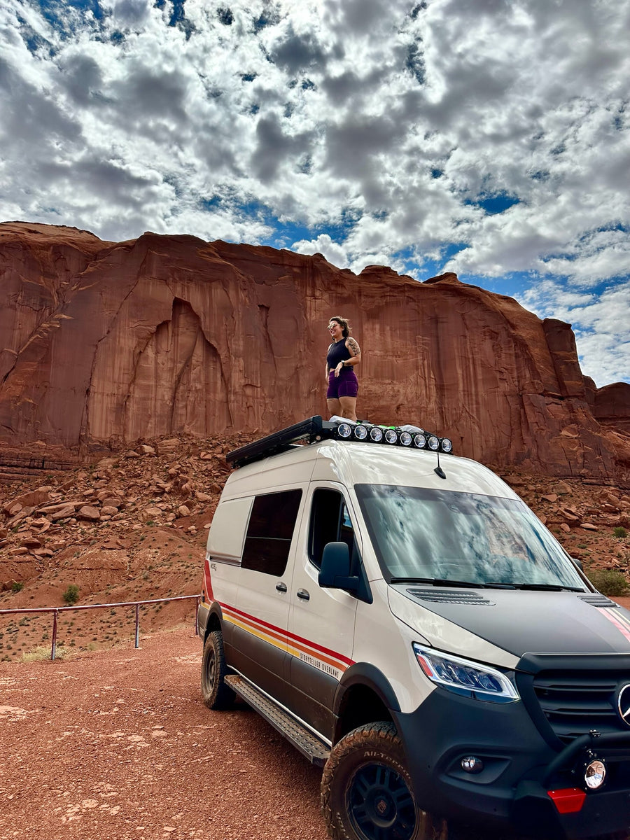 Woman on a Storyteller Overland RV rental van in a vast red rock canyon. Ideal for desert adventures!|White Storyteller Overland RV rental parked at a scenic campsite. A faint rainbow arcs over a rugged landscape with a grill nearby.|Storyteller Overland van