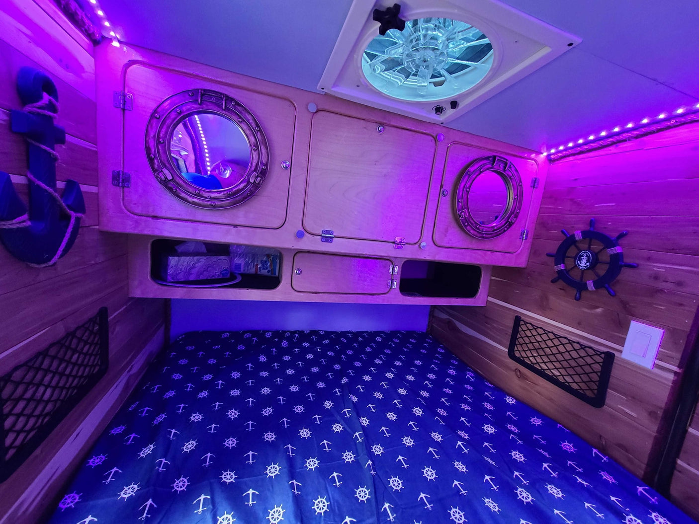 Nautical-themed RV rental interior. Purple LED lights highlight wooden walls, custom cabinets, bed with anchors, and a ceiling fan.