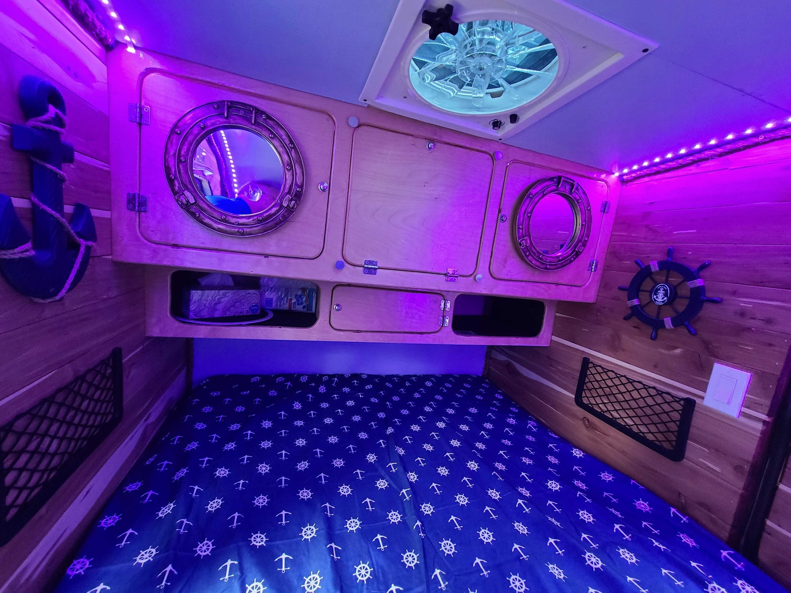 Nautical-themed RV rental interior. Purple LED lights highlight wooden walls, custom cabinets, bed with anchors, and a ceiling fan.