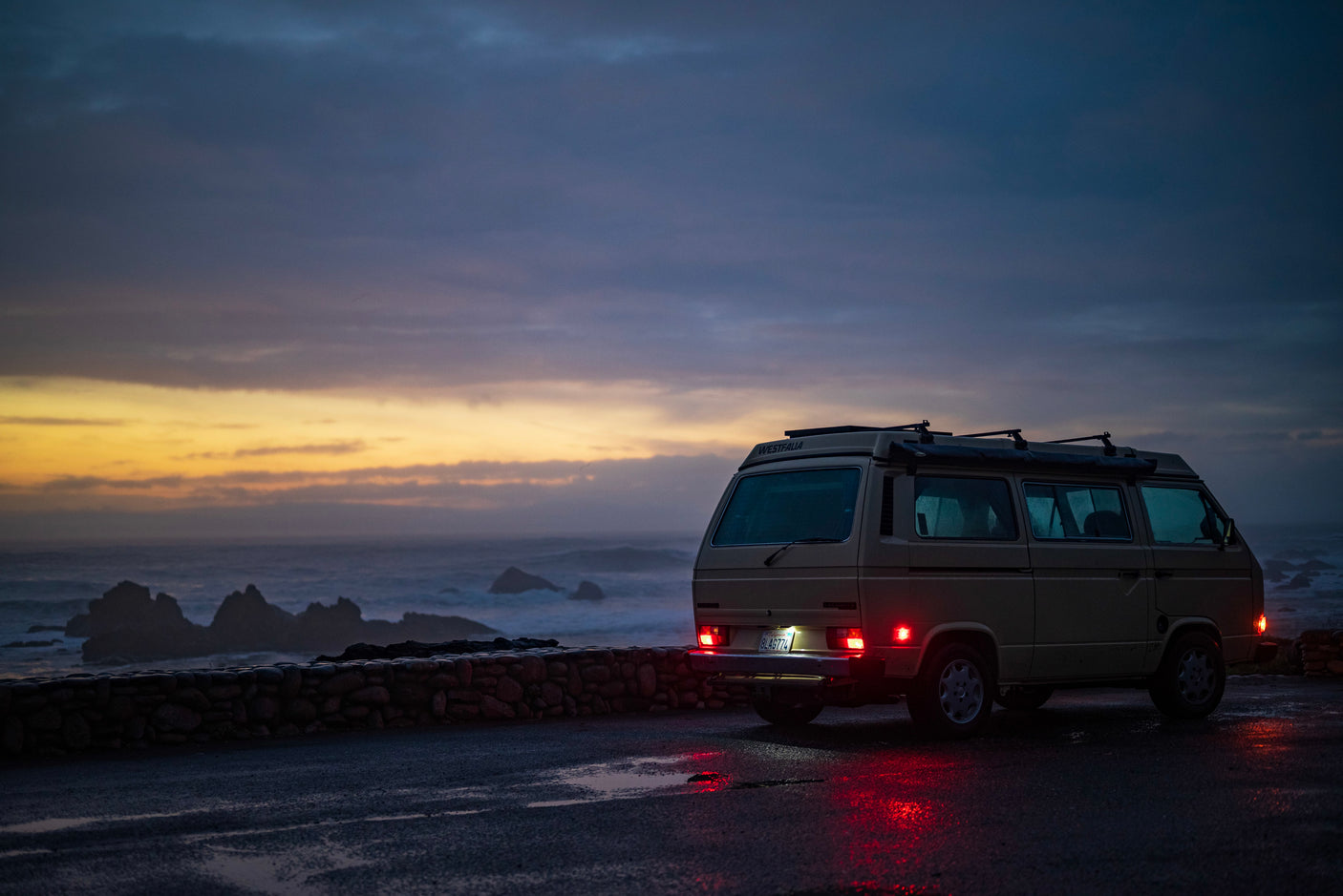Classic tan VW Westfalia camper RV rental by the ocean at sunset, red taillights reflecting on the wet road.