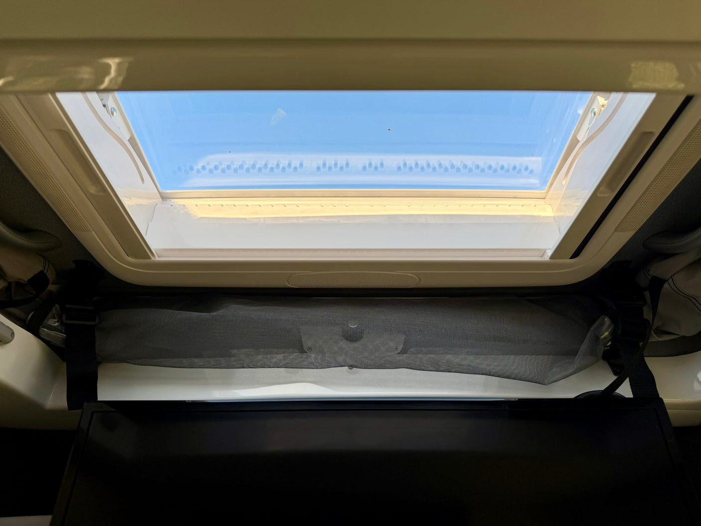 An RV rental's overhead skylight lets in blue sky. Below it are a retractable mesh screen and a dark privacy blind.