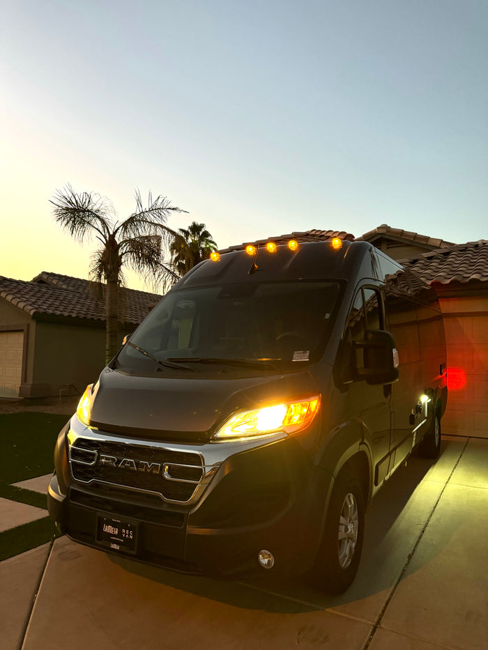 Dark grey Ram ProMaster RV rental van in a driveway at dusk. Headlights and amber roof lights are illuminated.