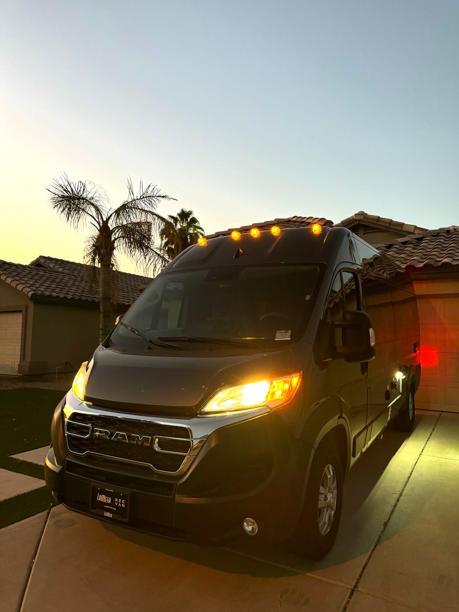 Dark grey Ram ProMaster RV rental van in a driveway at dusk. Headlights and amber roof lights are illuminated.