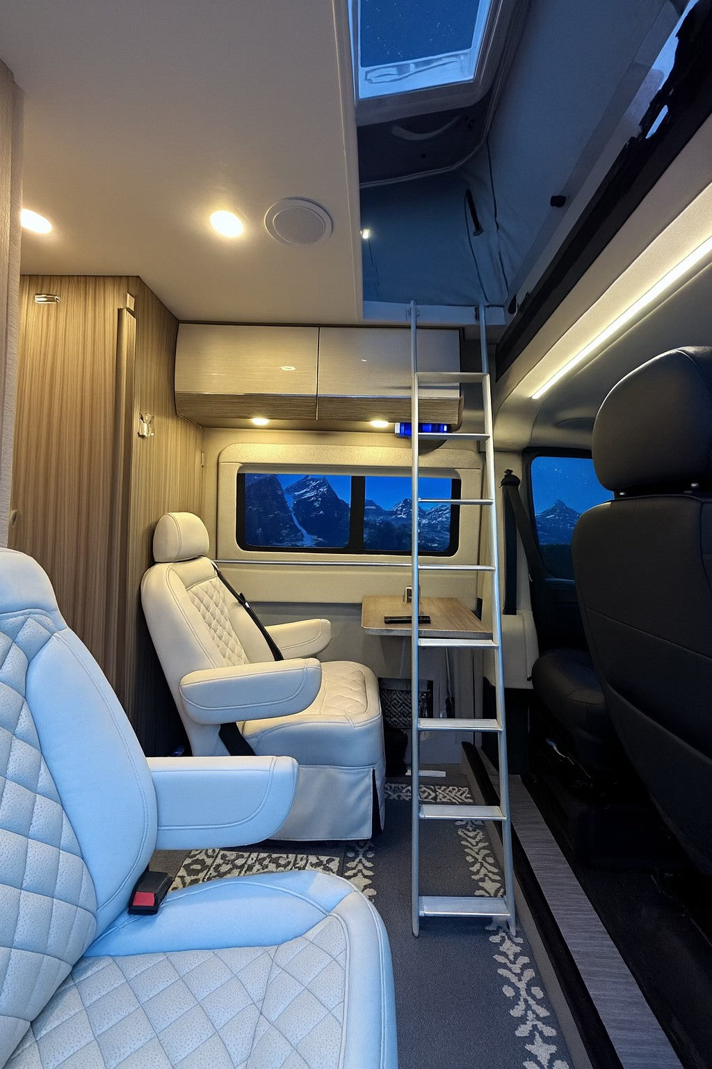 Luxurious RV rental interior featuring quilted white seating, a small table, ladder to a loft bed, and stunning mountain views.