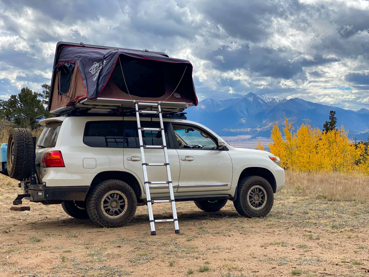 a child with blonde hair watches golden fall trees & mountains from a map-lined tent.|Enjoying a camping meal from this vehicle's setup by a scenic lake. Like an RV rental