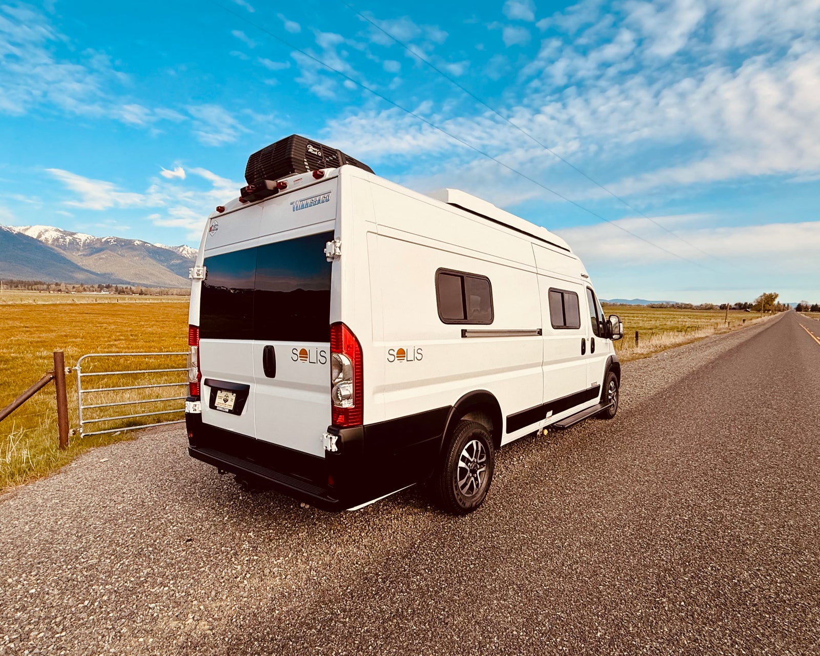 This white Solis rv rental van offers adventure. Parked roadside with mountains & fields, ready for camping & scenic drives.