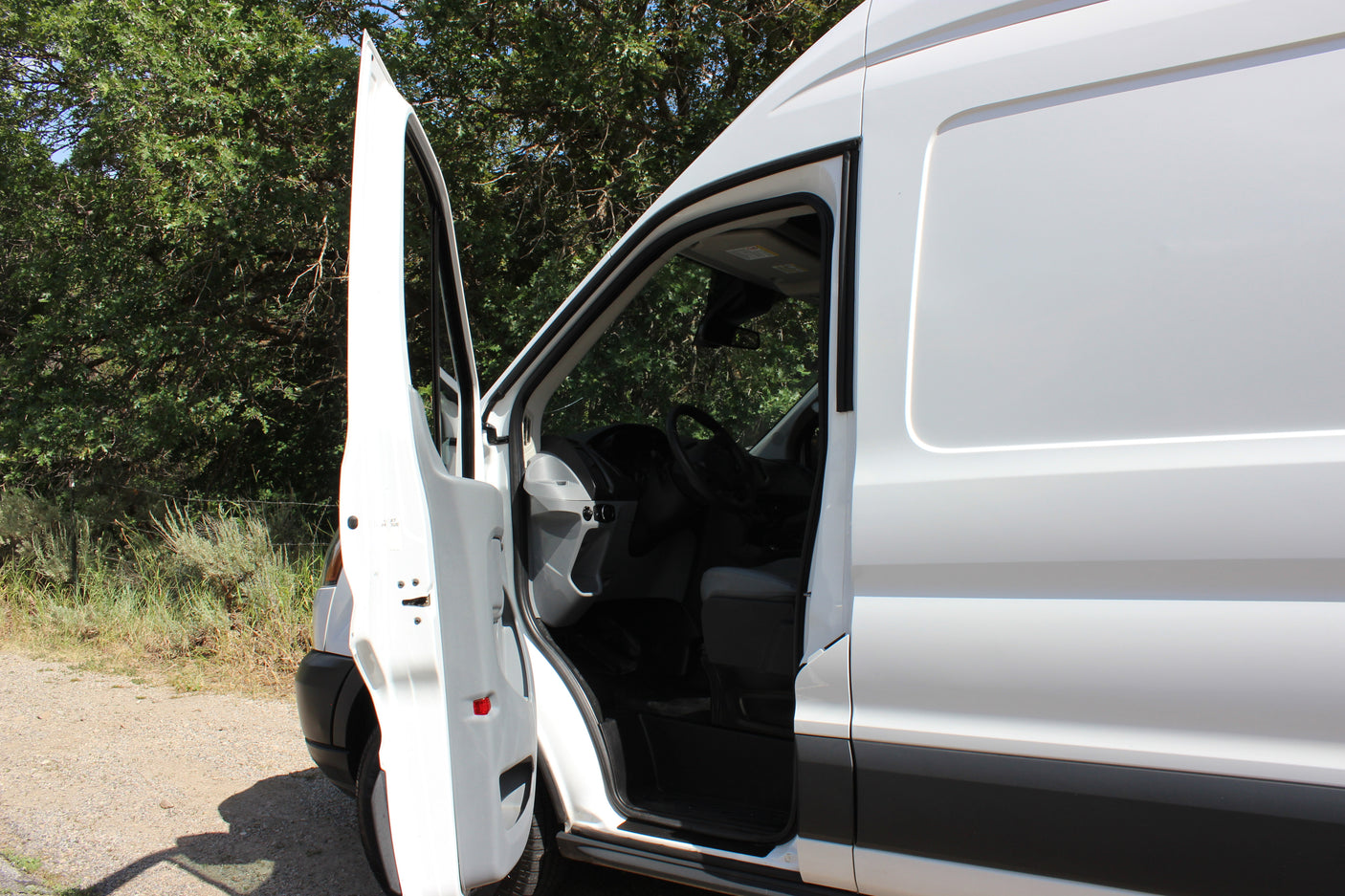 This white RV rental van has its driver's door open, revealing the interior cabin, parked on a gravel path near green trees.