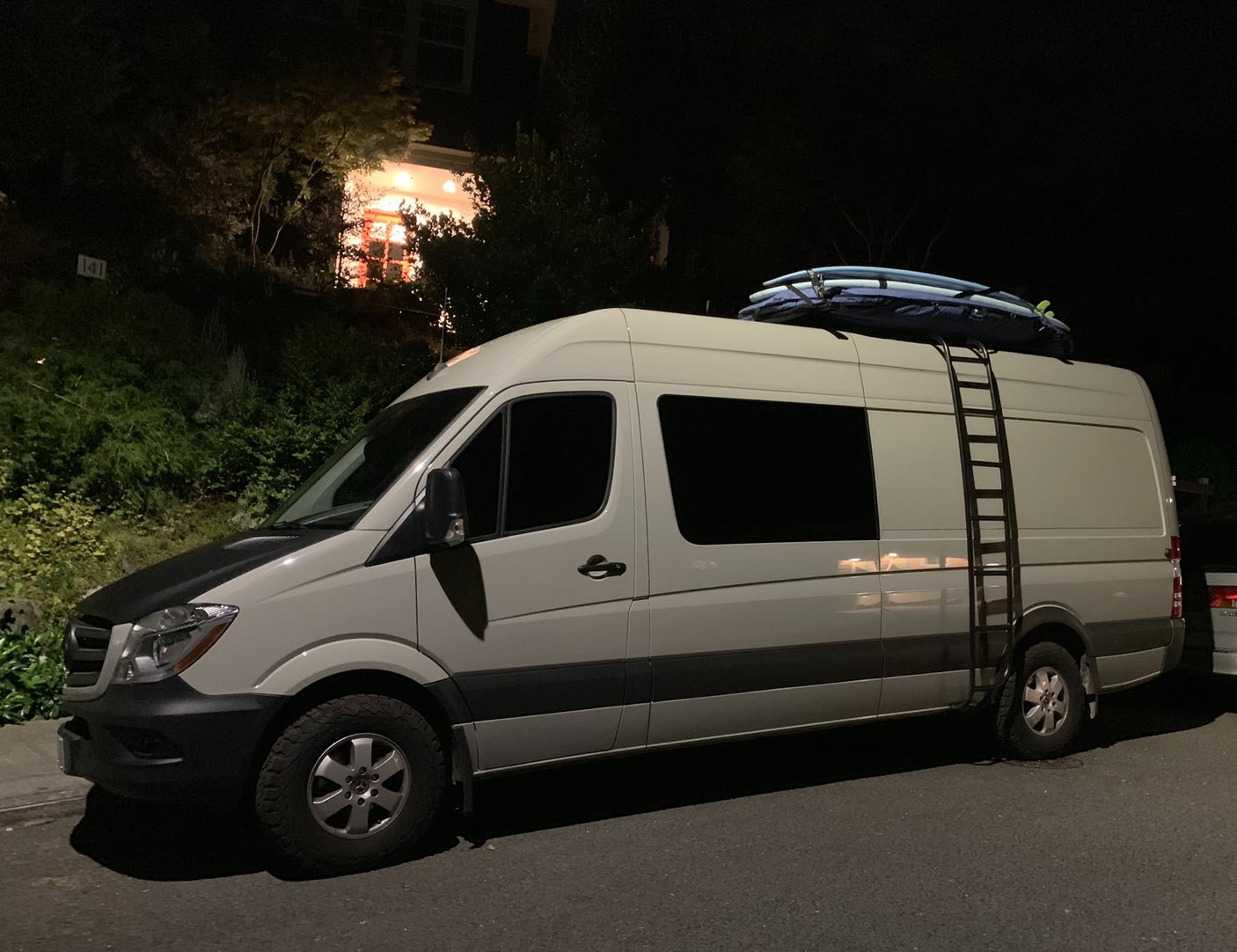 A white RV rental van, topped with surfboards and a ladder, parked on a residential street at night.