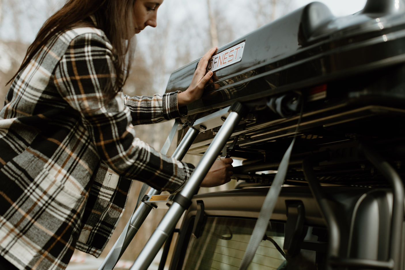 A woman in a plaid shirt prepares a black OFNEST rooftop tent on a vehicle, adjusting straps for an RV rental adventure.