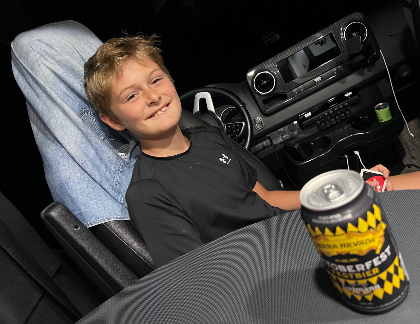 A smiling child in a modern RV rental, an adult's legs up, with a Sierra Nevada beer can & playing cards nearby.