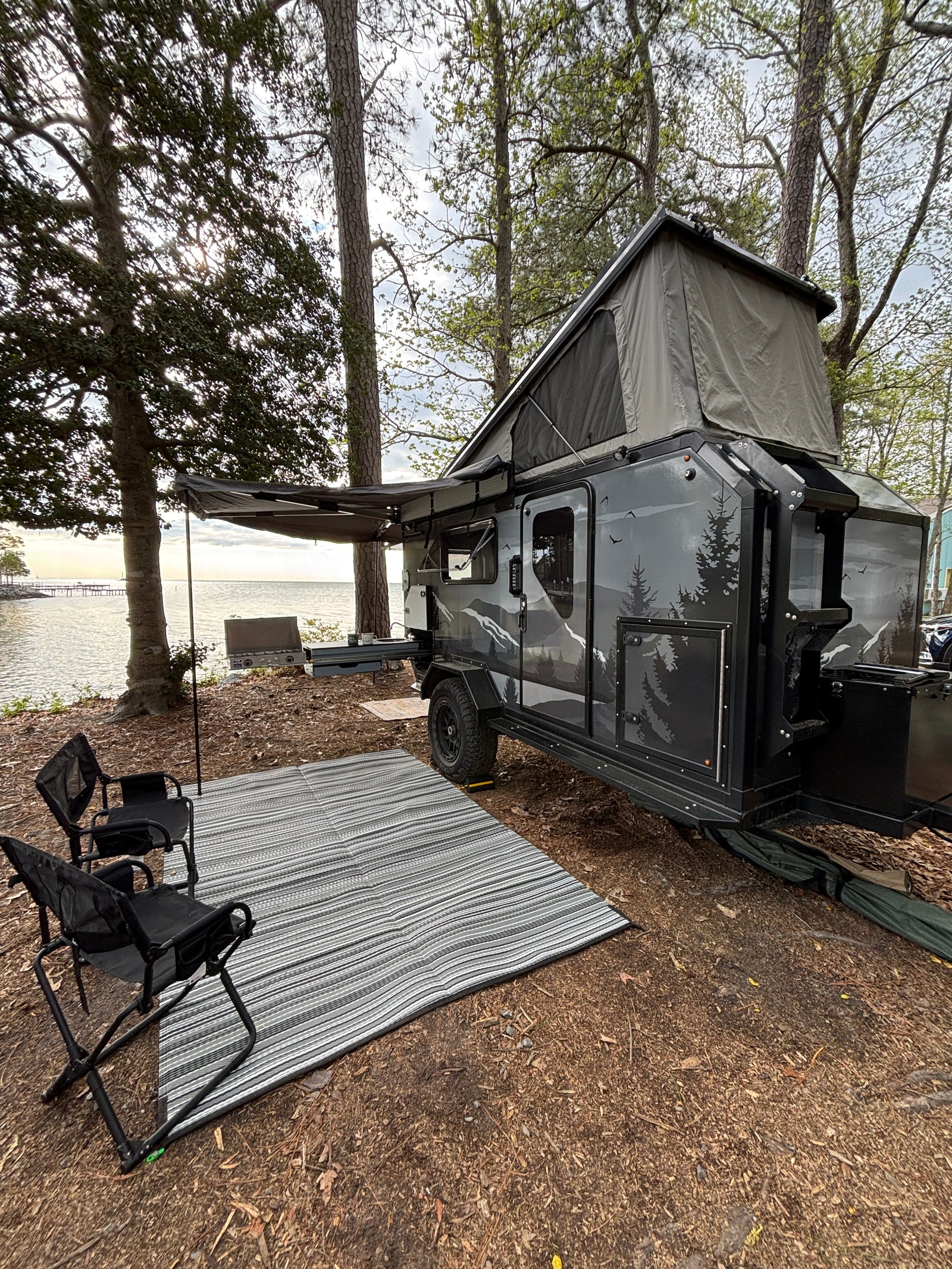 This RV rental features a pop-up camper trailer by the water with an awning, outdoor mat, and camp kitchen setup.