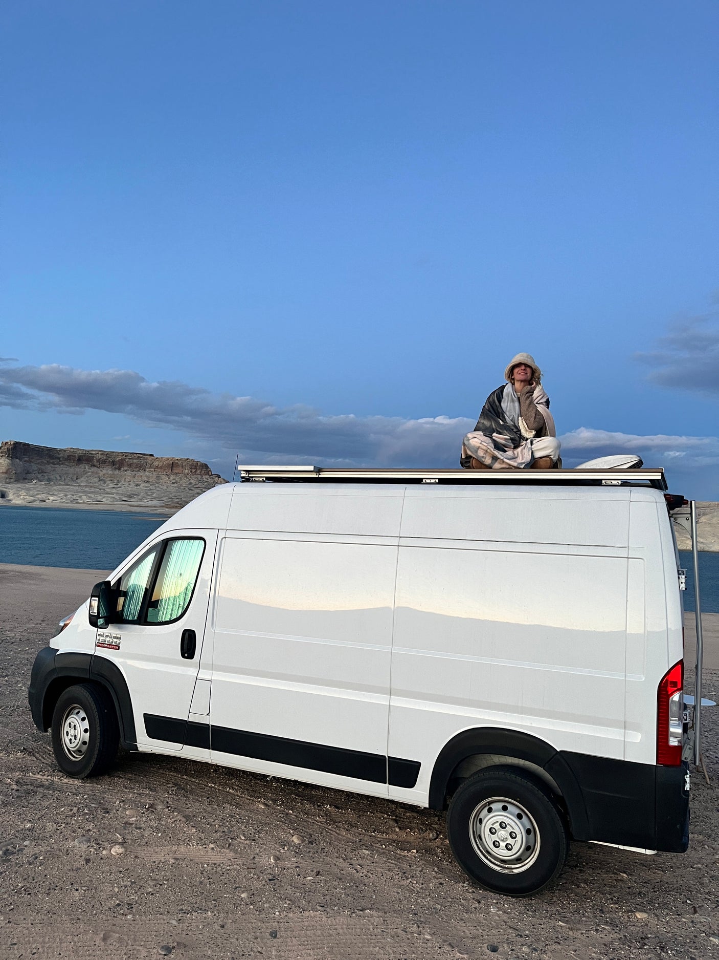 Cozy moment on an RV rental van's roof overlooking a lake. A person in a blanket enjoying their van life adventure.