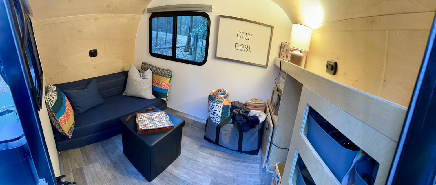Cozy RV rental interior: sofa, Scrabble, "Our Nest" sign, and nature views through the window.