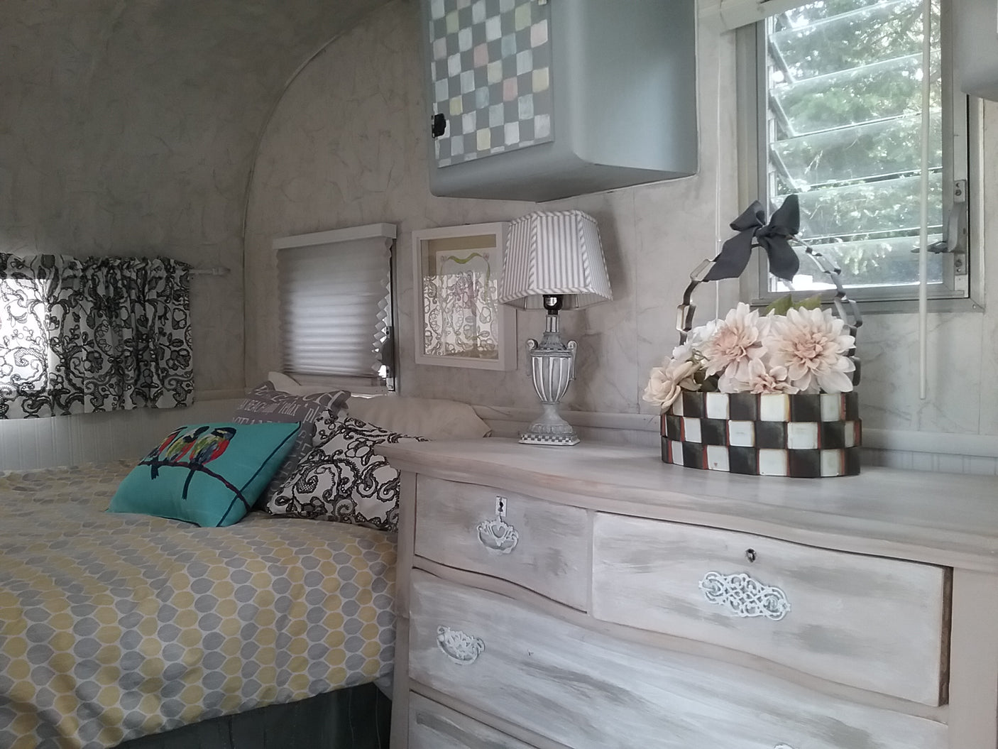 Inside a charming RV rental: a comfy bed with patterned bedding, a distressed dresser holding a lamp and floral decor.