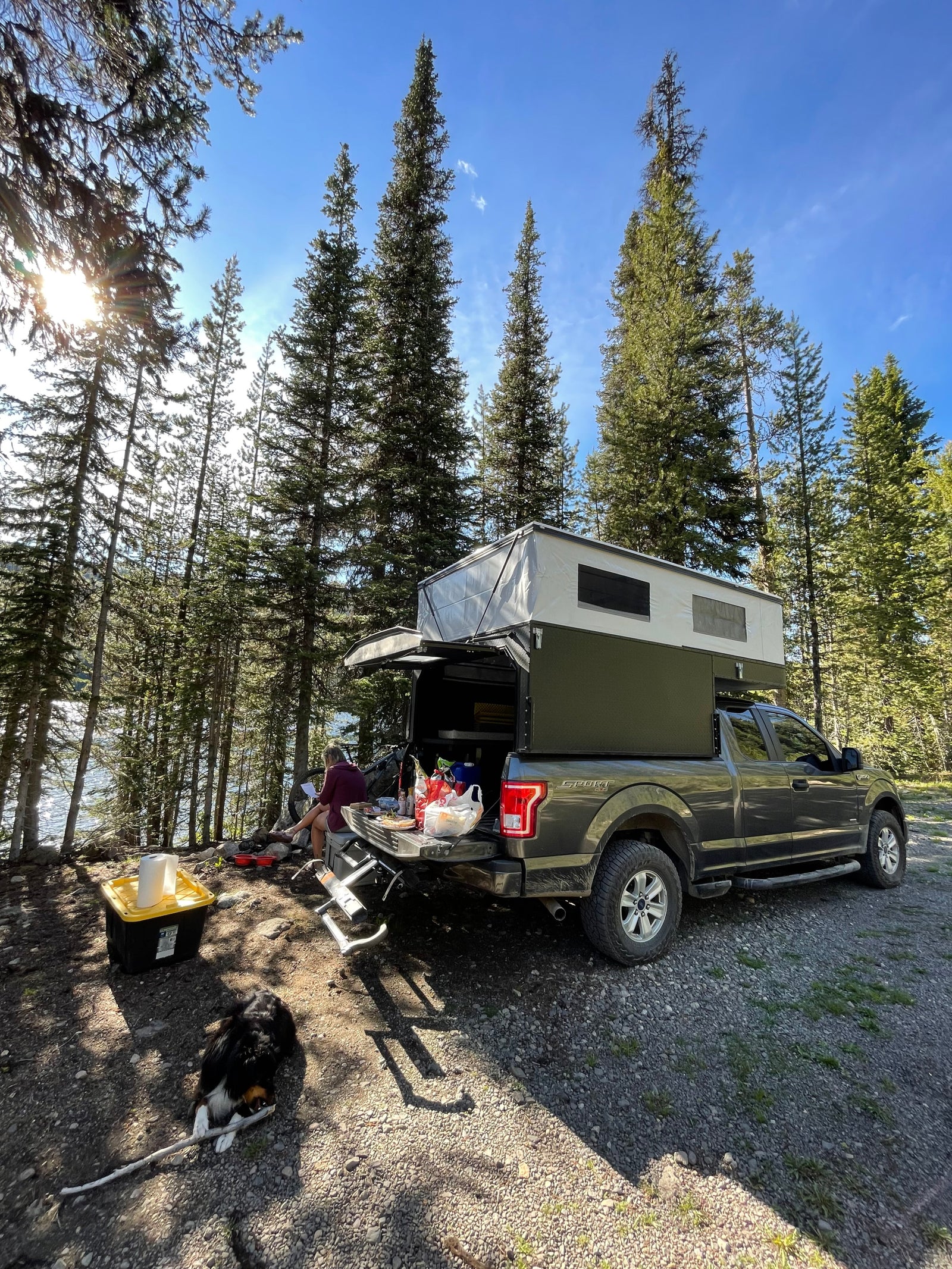 A person and dog relax by a lake next to a pop-top truck camper. Explore nature with an RV rental like this!