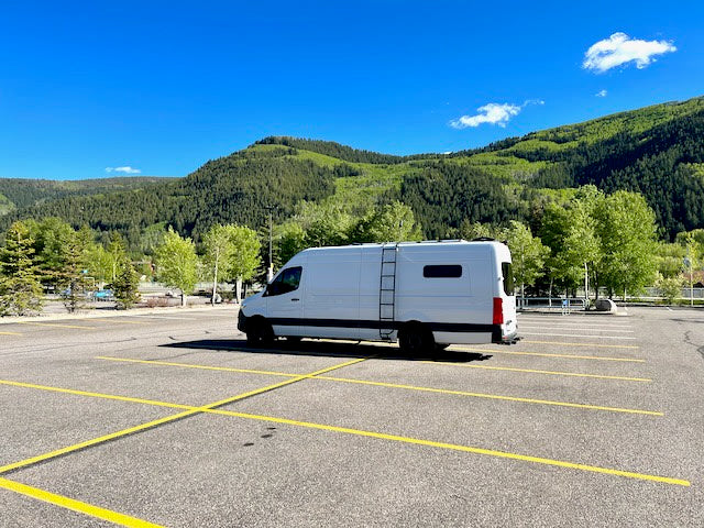 White RV rental van parked alone in a scenic mountain lot. Blue sky & green trees enhance the travel vibe.
