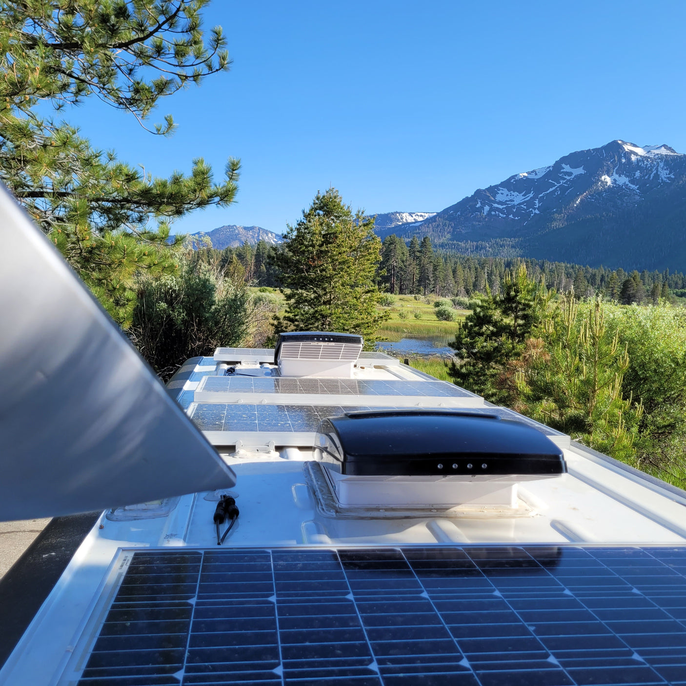 Looking out from an RV rental, showing solar panels on the roof, with a serene view of mountains, a lake, and pine trees.