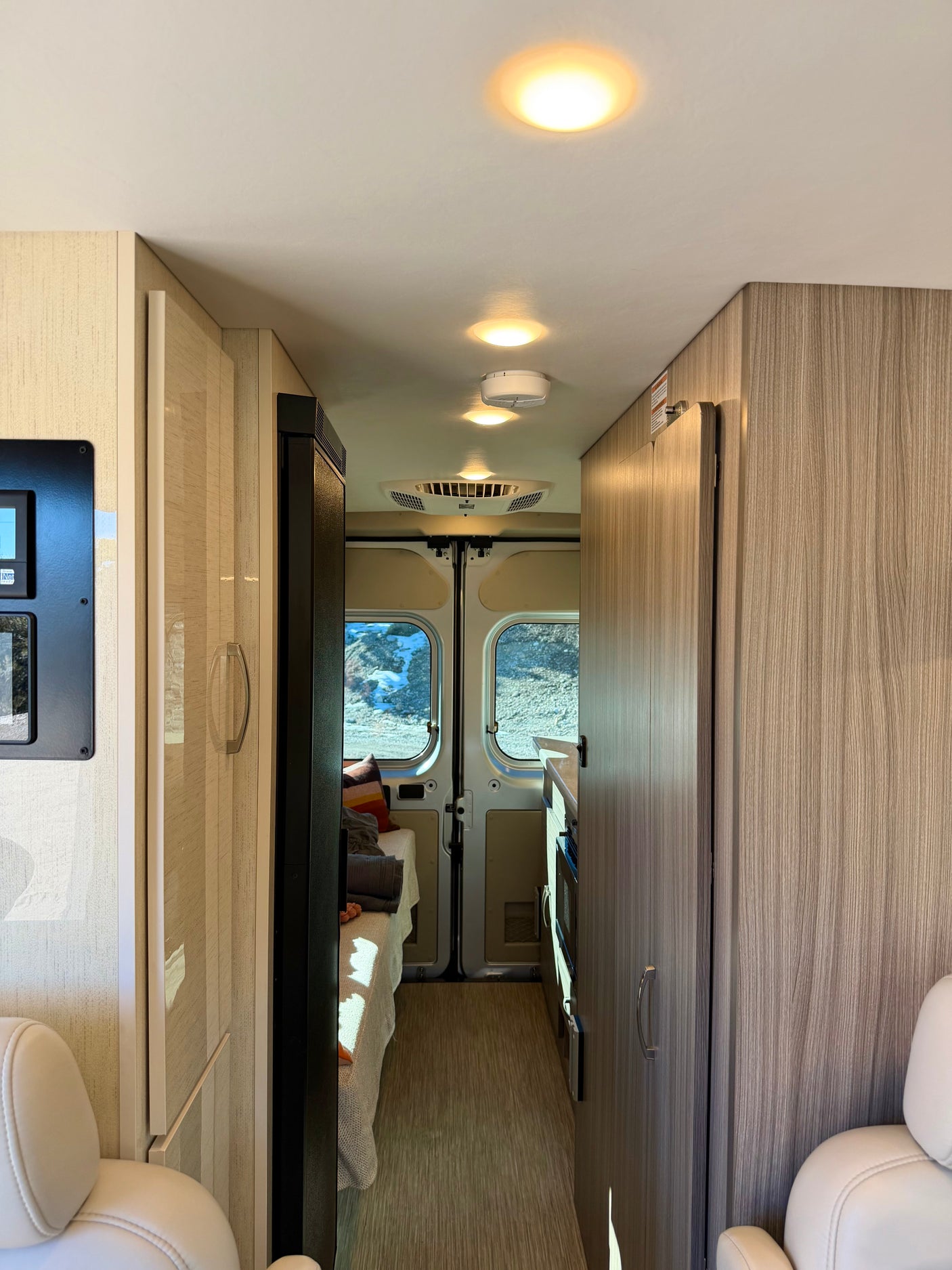 Cozy rv rental interior, looking past front seats to kitchen, bed, and rear windows. Light wood cabinetry & bright lights.