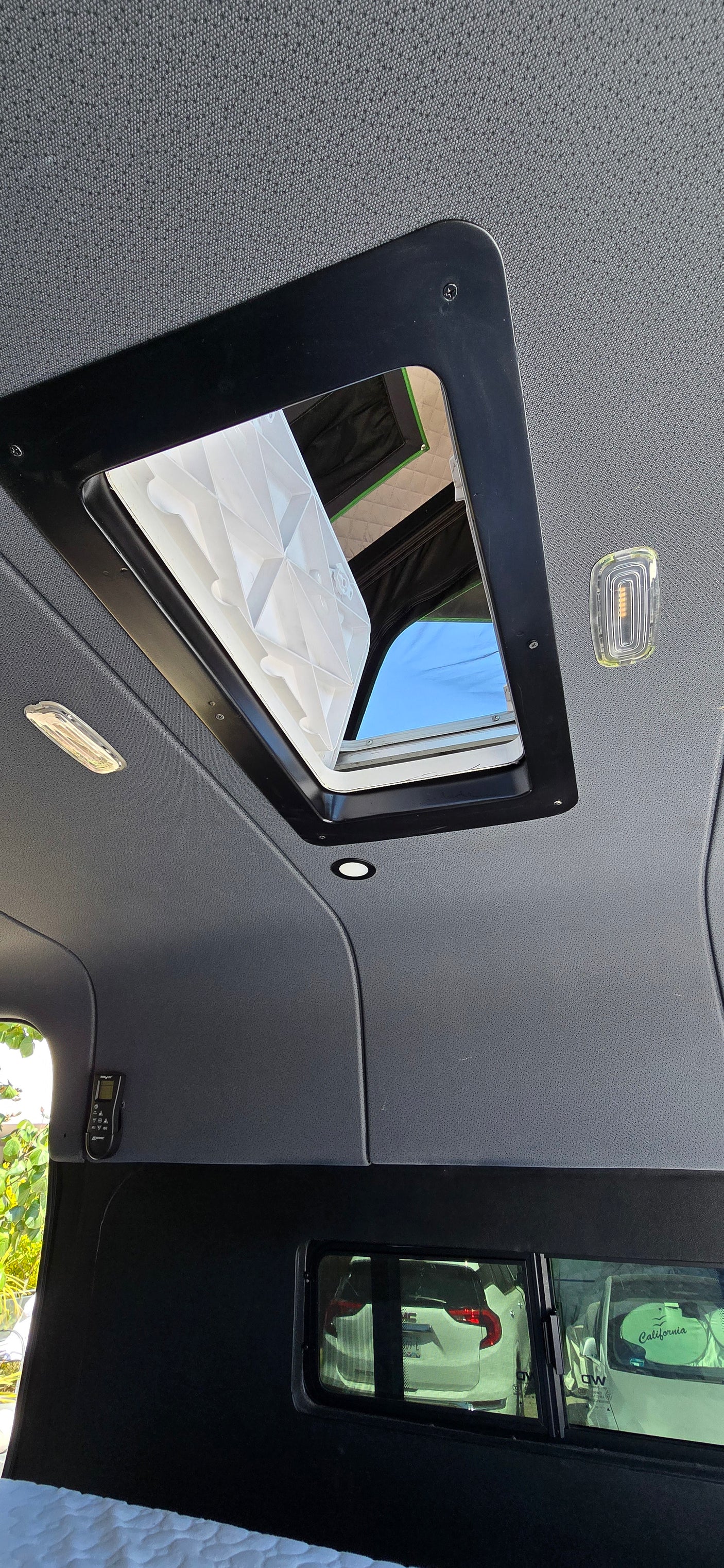 Interior of an RV rental: An open roof vent, overhead lights, a wall-mounted control panel, and a side window view.