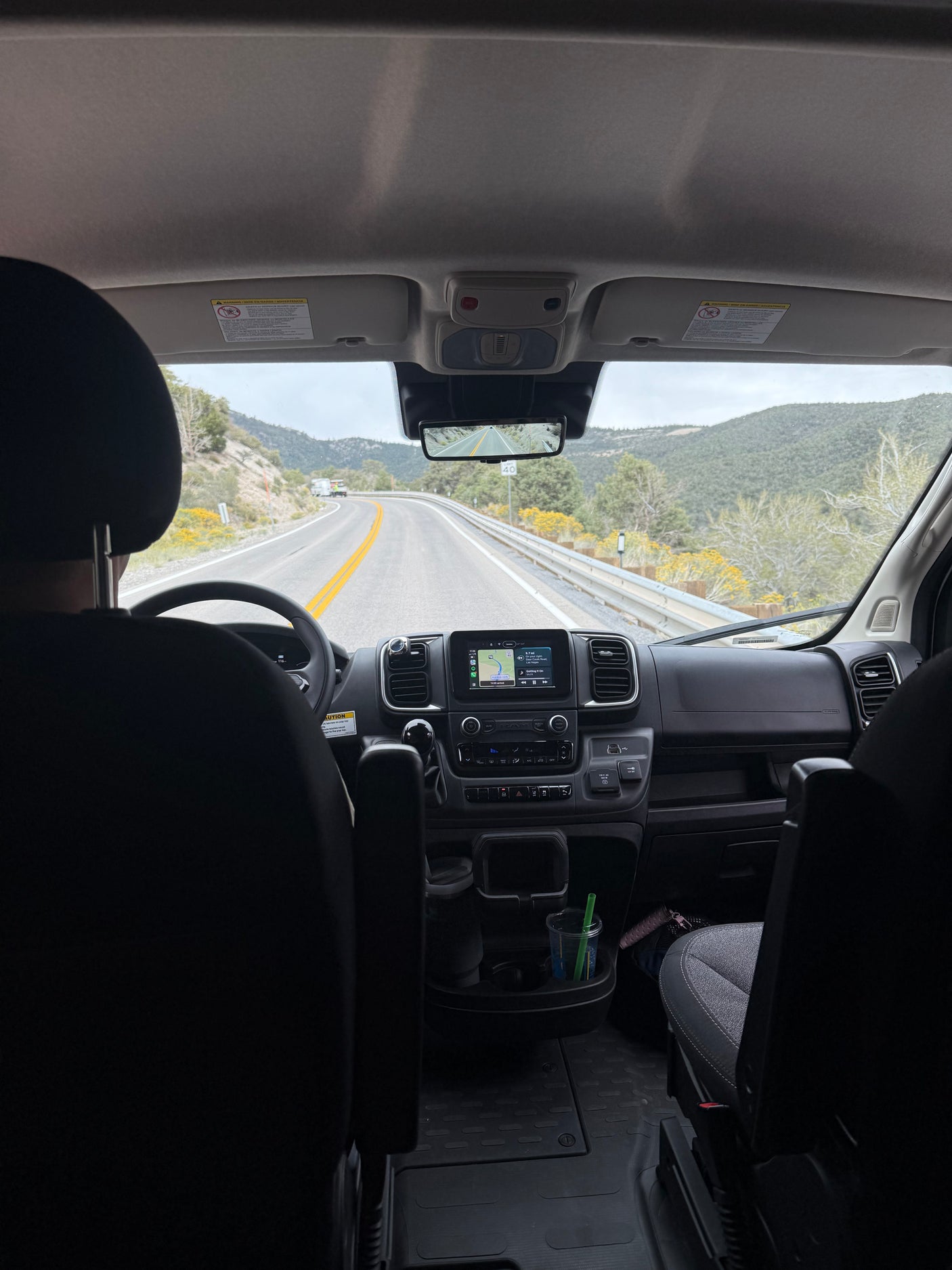View from an RV rental's passenger seat. Driver steers on a mountain road with navigation active, cloudy skies above.