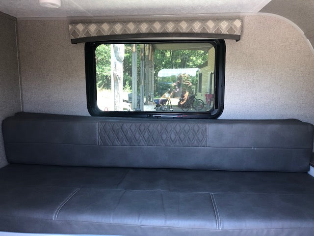 RV rental interior: comfy grey dinette/bed and window reflecting a person, bike, and trees outside.