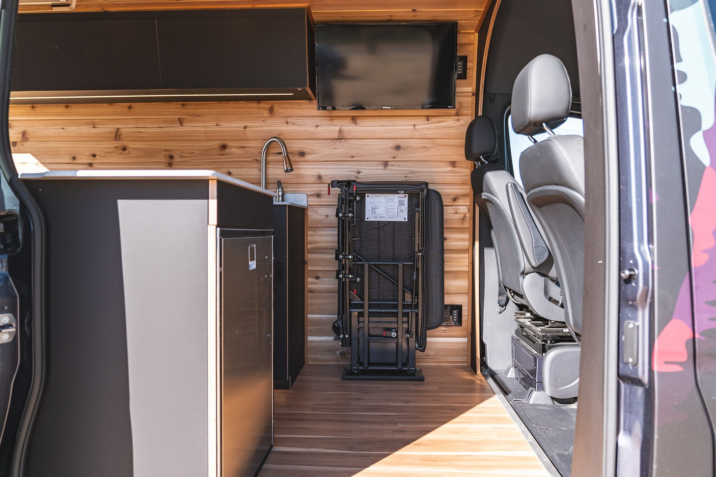 Modern RV rental interior with warm wood paneling, compact kitchen area including sink and fridge, plus foldable passenger seating.