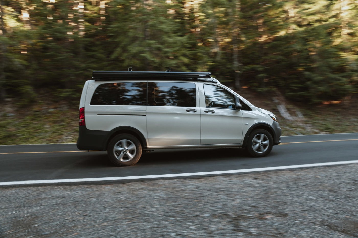 parked by a lake. Surrounded by mountains & evergreen trees under cloudy skies.|RV rental: Light campervan with roof awning on a blurred forest road — photo 2