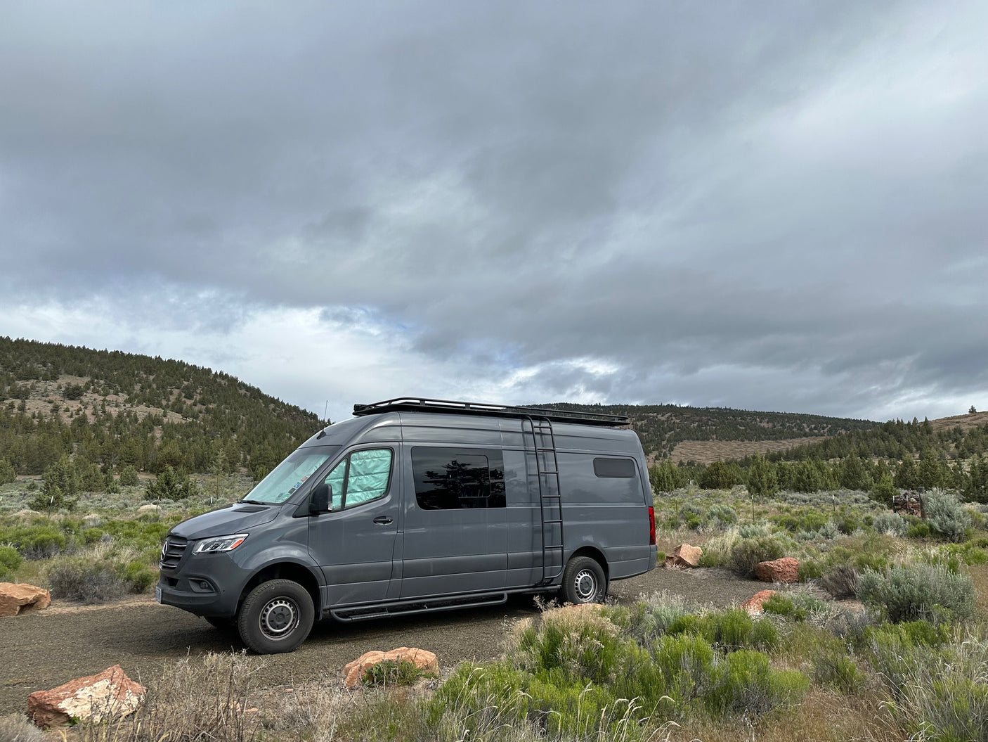 Gray RV rental camper van parked on a dirt road, ready for a road trip adventure through hills & shrubs under cloudy skies.