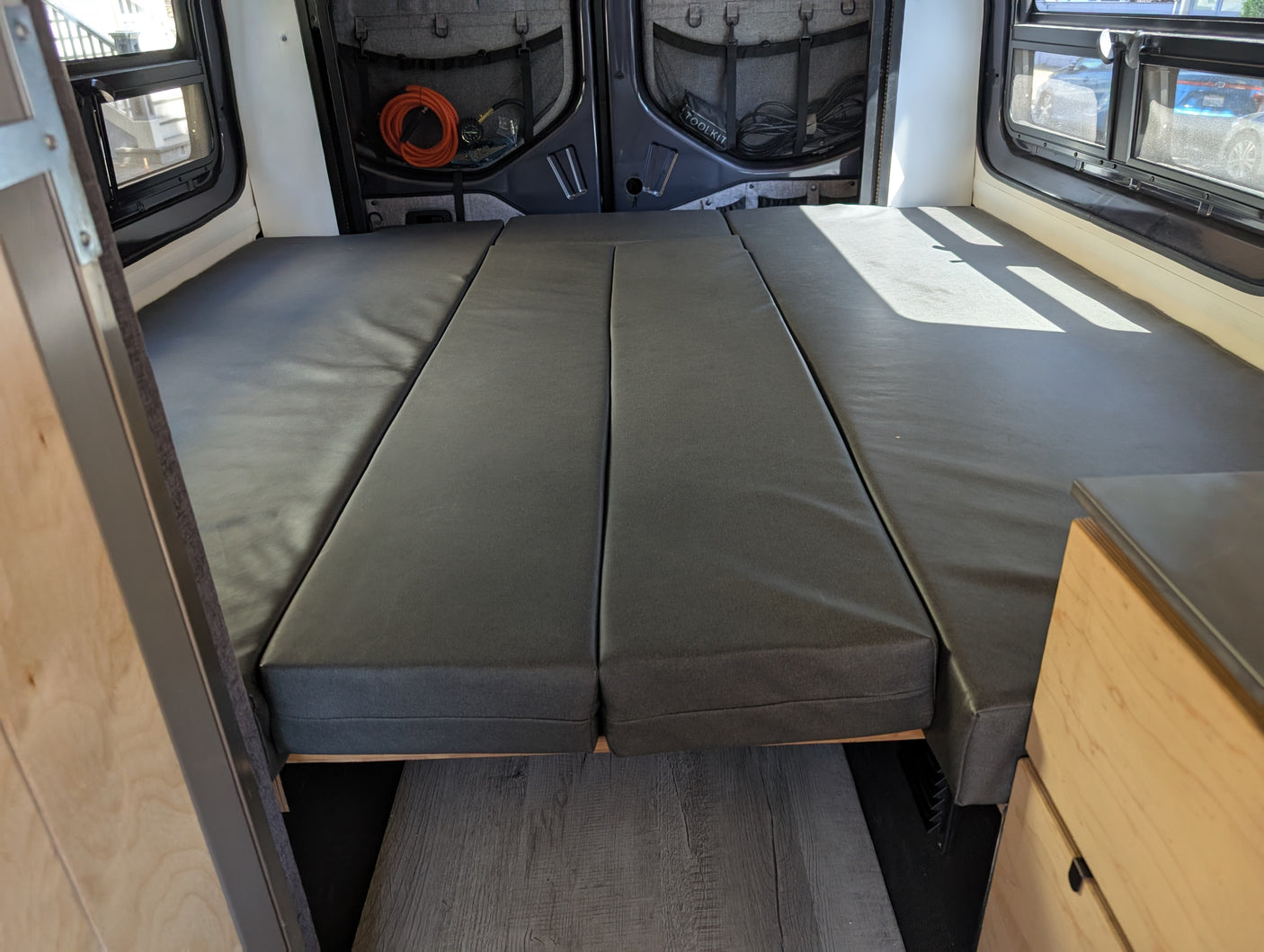 RV rental: Camper van interior with a grey modular bed, rear windows offering natural light, and organized storage on back doors.