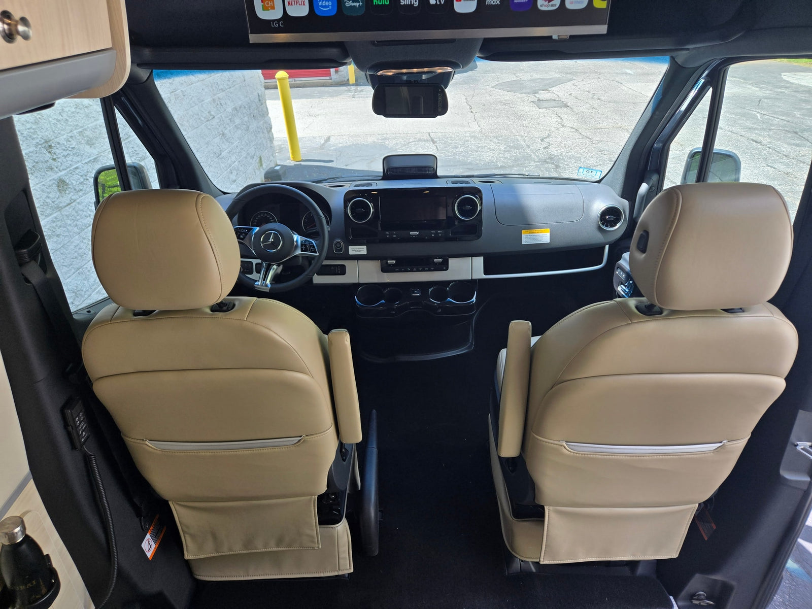 Interior of an RV rental: beige front seats, Mercedes-Benz cockpit, and dashboard with infotainment. Overhead screen shows apps.