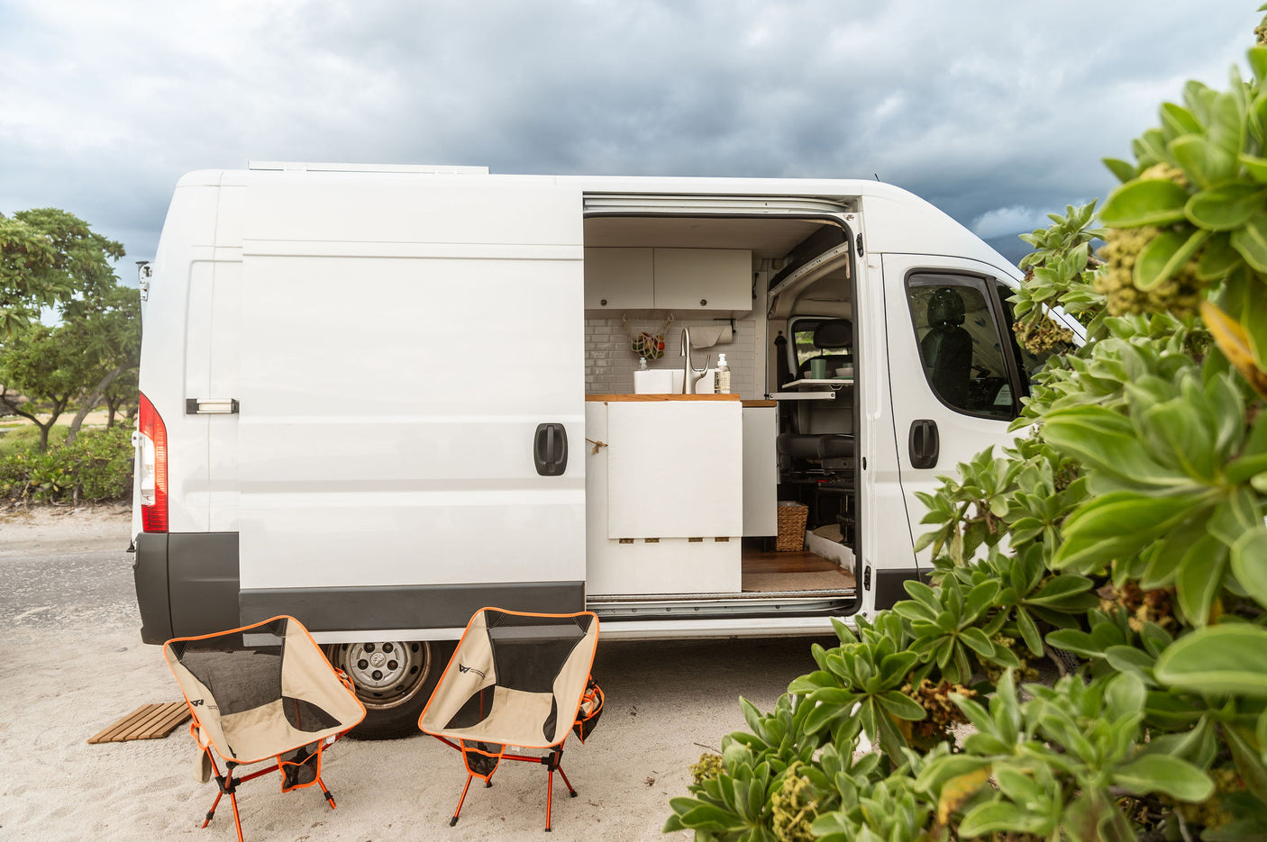 RV rental camper van, side open, shows modern kitchen & outdoor chairs. Parked in nature, ready for travel adventures.