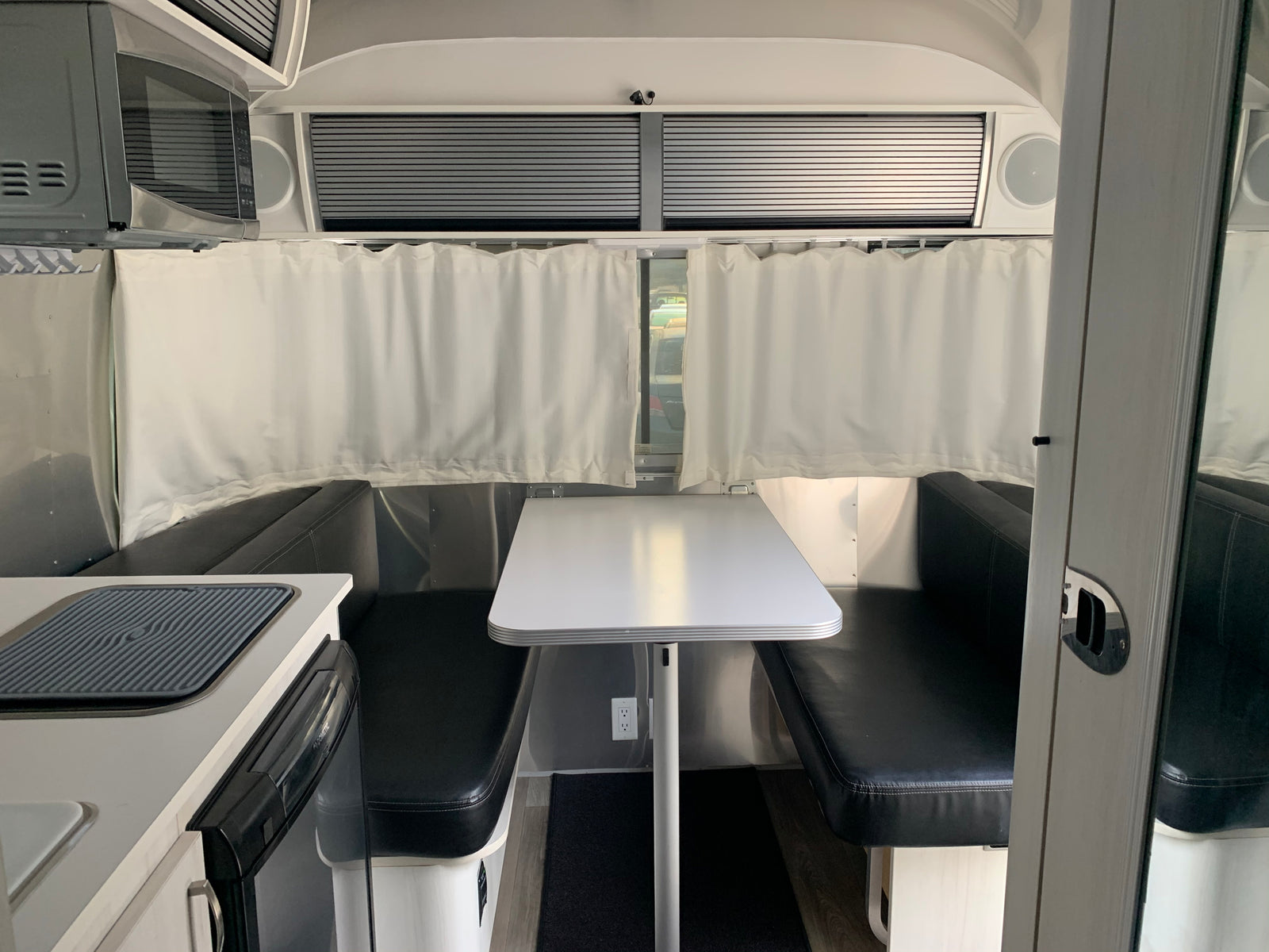 Modern RV rental interior, showing a dining area with black benches, white table, microwave, and kitchen.