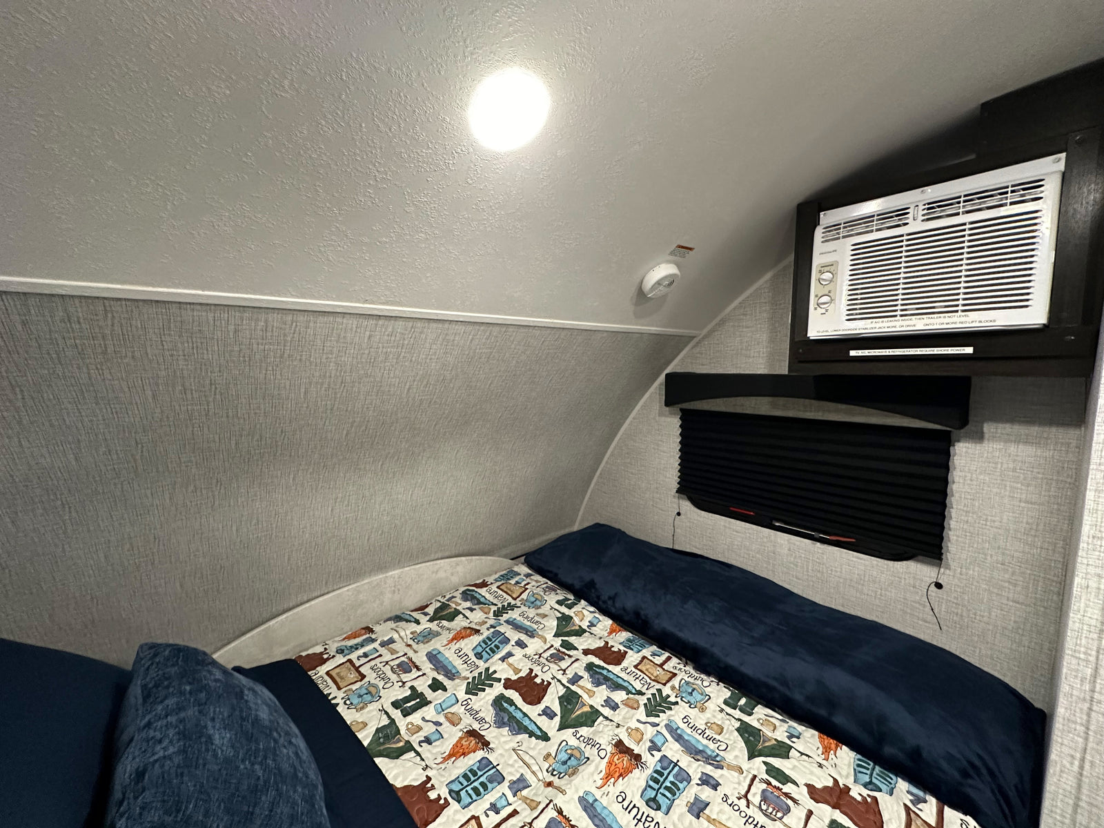 Cozy RV rental bedroom featuring a bed with a camping-themed blanket, wall-mounted AC unit, and smoke detector.