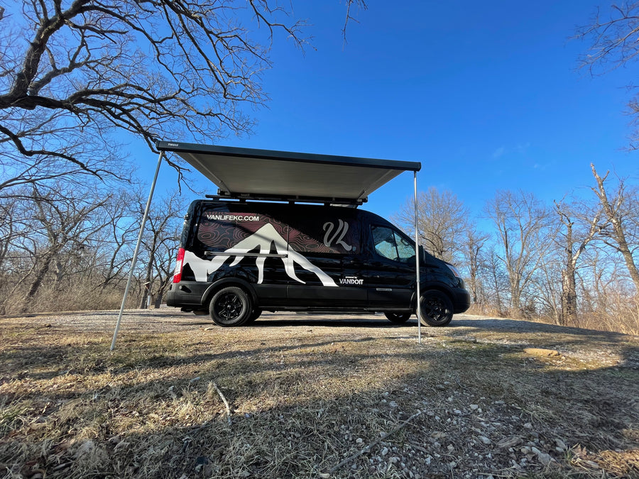 This black **rv rental** camper van, awning out, sits in a dry wooded area under a clear blue sky.