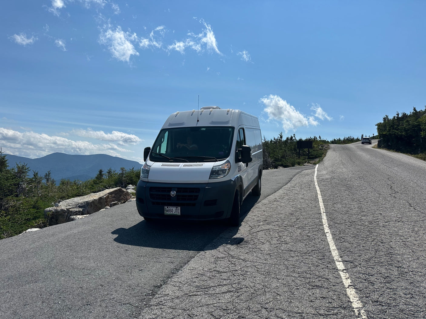 A white RV rental camper van is parked on a scenic mountain road under a bright blue sky, with distant peaks visible.
