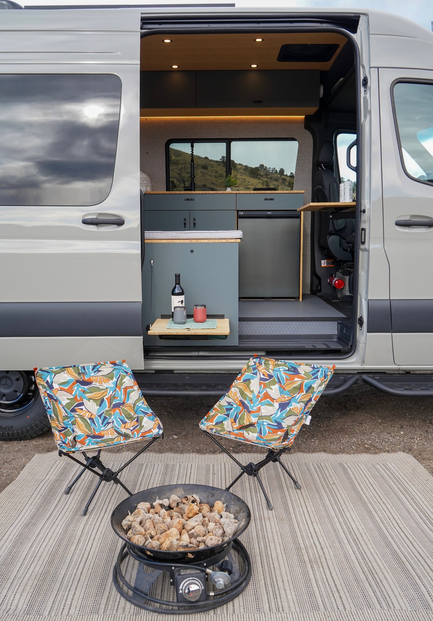 RV rental setup: A camper van with its door open, showing a kitchen, two outdoor chairs, and a fire pit on a rug.