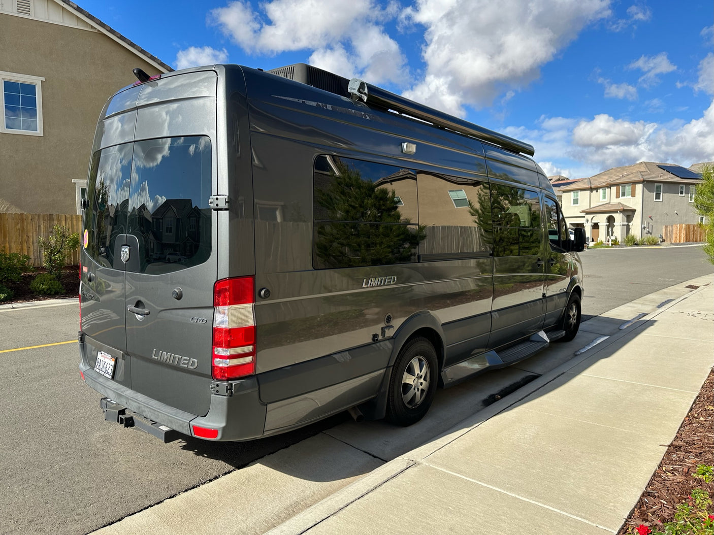 Stylish gray camper van, perfect for an RV rental, parked on a sunny residential street. Features include roof AC and awning.
