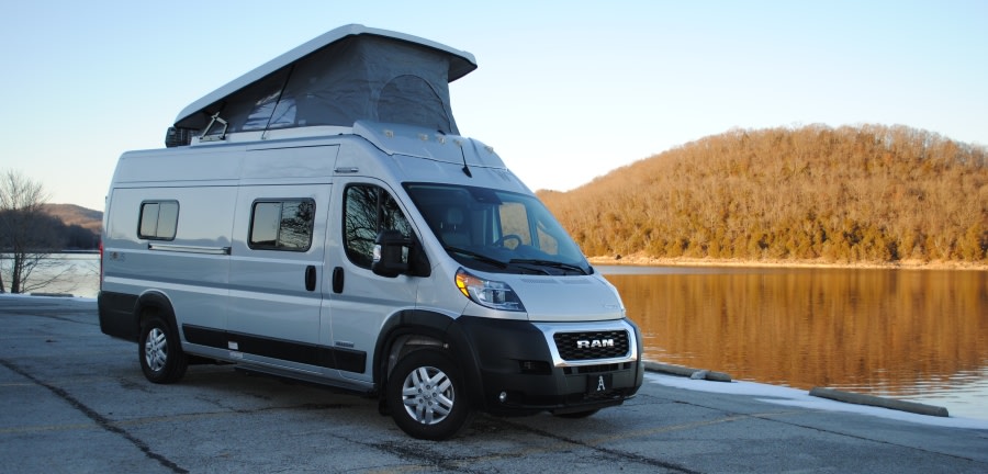 parked by a calm lake with hills beyond.|Explore the outdoors! A silver Ram ProMaster pop-top camper van