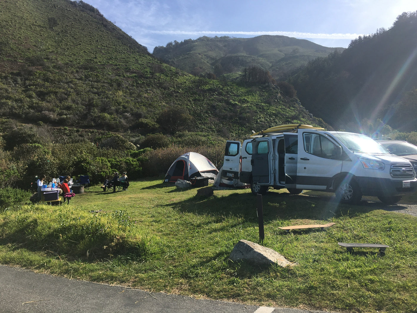 RV rental camper van and tent at a sunny campsite with people, nestled in lush green hills.