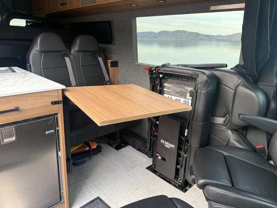 This RV rental interior features comfortable seating, a versatile table, an Isotherm fridge, and a stunning lake view.