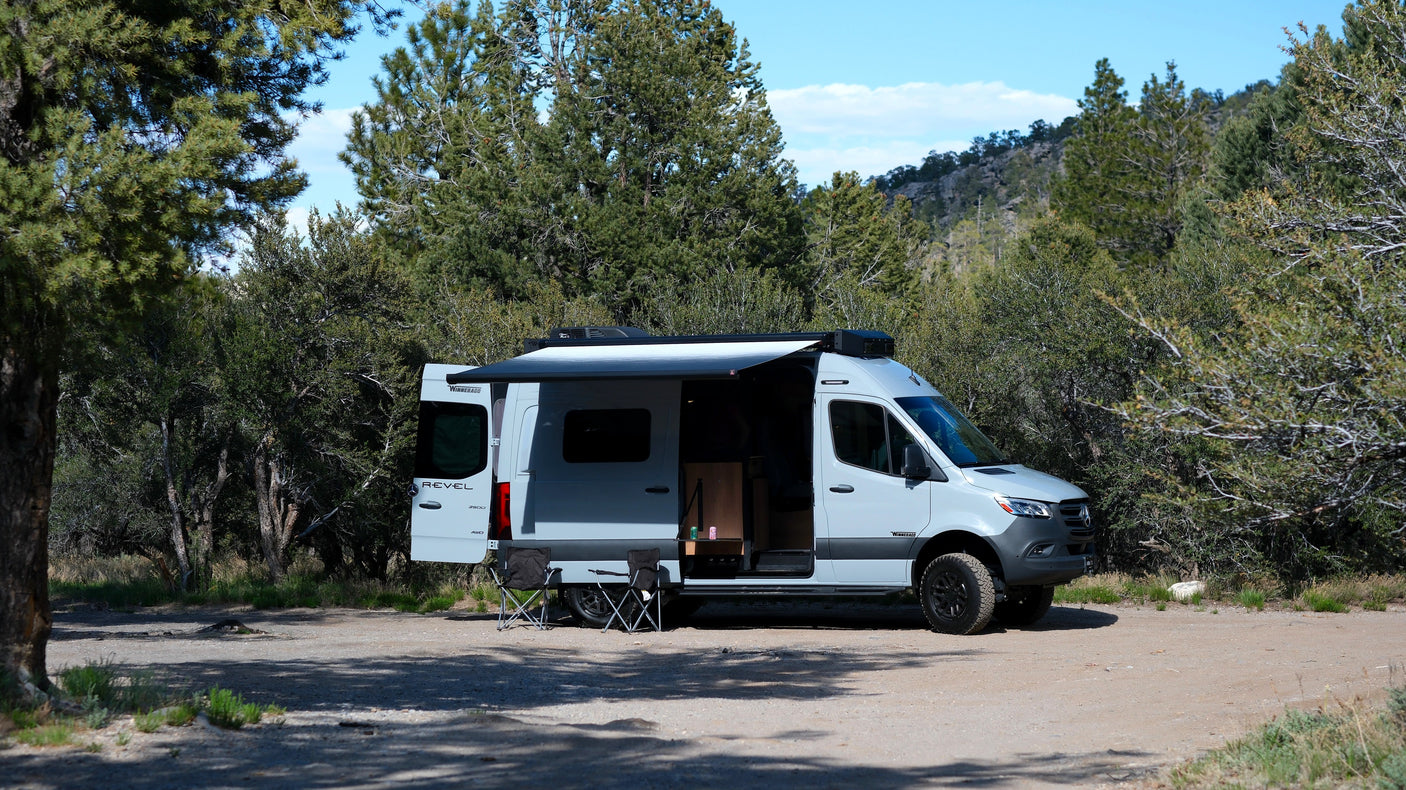 Light grey Winnebago Revel rv rental camper van parked in a wooded area, awning out, chairs ready for camping adventure.