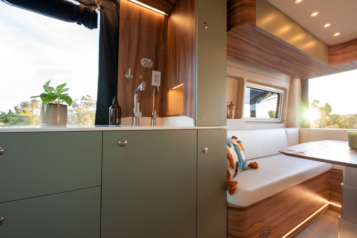 Modern RV rental kitchen and dining area. Olive green cabinets, wood accents, and bright windows create a cozy camper interior.